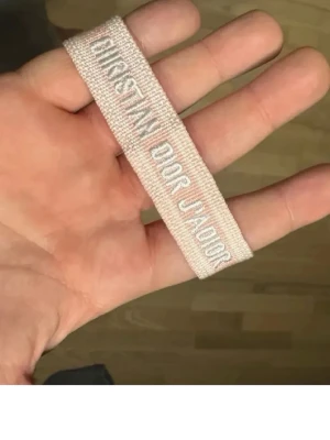 Christian Dior rosa tygarmband - Säljer ett ljusrosa tygarmband från Christian Dior med texten 'Christian Dior J'Adior' broderat i vitt. Armbandet har justerbara knytband och en stilren, trendig look som passar perfekt till din outfit.