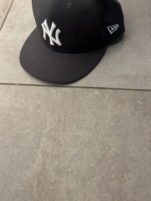 New Era NY keps mörk grå - Snygg grå keps från New Era med klassisk New York Yankees-logga i vitt framtill. Kepsen har rak skärm och broderad logga på sidan. Tillverkad i slitstarkt tyg, perfekt för en streetwear-look.