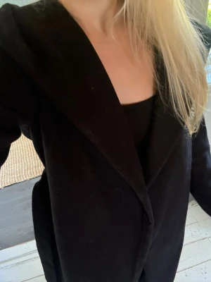 Svart lång kappa med skärp - Stilren svart lång kappa med huva och knytband i midjan. Kappan är ifrån Chelsea