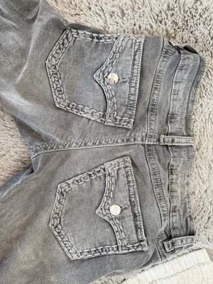 Grå jeans med detaljerade bakfickor - Säljer ett par grå jeans med unika bakfickor som har dekorativa sömmar och silverfärgade knappar. Jeansen har låg midja, bälteshällor och klassisk femficksdesign. Materialet är denim i bomull och passformen är bootcut. Från Gina i storlek 158.