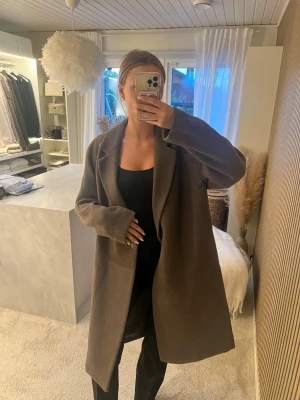 Brun oversized kappa - Snygg brun oversized kappa i ullmix med klassisk krage och raka ärmar. Kappan har en enkel, stilren design utan synliga knappar och går ner till knäna. Perfekt för att hålla värmen under kyliga dagar och passar till många olika outfits.