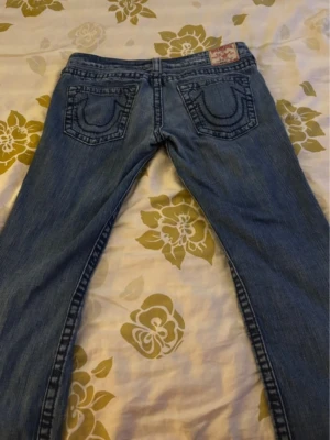Blå jeans med broderade bakfickor - Säljer ett par klassiska blå jeans med snygga broderade detaljer på bakfickorna och låg midja. Jeansen har raka ben och slitningar framtill för en cool look. Materialet är denim i bomull och de har knappgylf samt nitar vid fickorna.