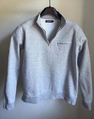 Ljusgrå halvzip sweatshirt, Dressman - En stilren ljusgrå sweatshirt med halv dragkedja och liten bröstficka. Tröjan har ribbade muddar vid ärmslut och nederkant samt en hög krage som kan bäras öppen eller stängd. Perfekt för en avslappnad och trendig look.