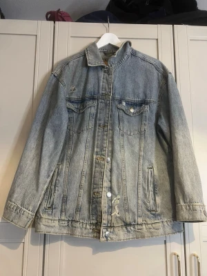 Oversized jeansjacka från Stradivarius - Ljusblå oversized jeansjacka från Stradivarius med slitna detaljer och klassisk knappstängning. Två bröstfickor med lock och knappar samt två sidofickor. Perfekt för en avslappnad och trendig look.