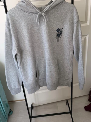 Grå hoodie med blå ros-tryck - Snygg grå hoodie med huva och dragsko. På bröstet finns ett blått ros-tryck och på ryggen ett större tryck med rosor och texten 'Nature Blossom'. Hoodien har en stor magficka och ribbade muddar vid ärmar och nederkant. Perfekt för en chill och trendig look.
