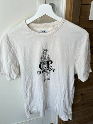 Vit t-shirt från C.P. Company - Vit t-shirt från C.P. Company med tryck av en figur och logga på bröstet. Klassisk rund hals och korta ärmar. Tillverkad i mjuk bomull som känns skön mot huden. Perfekt för en avslappnad och stilren look.