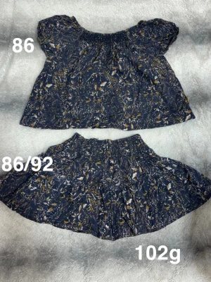 Blommigt set med topp och kjol - Sött set med en mörkblå topp och matchande kjol, båda med  mönster i vitt och gult. Toppen har korta puffärmar och resår vid halsen, kjolen har resår i midjan för skön passform. Perfekt för dig som gillar lekfulla och bekväma plagg.🌸Fint använt skick. Storlek 86 på tröjan och 86/92 på kjolen. 