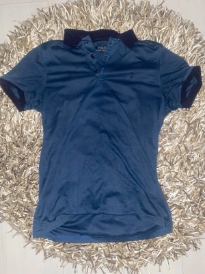 Blå piké t-shirt från Polo Ralph Lauren - Blå piké t-shirt från Polo Ralph Lauren med mörkblå krage och ärmslut. Klassisk design med knappar vid halsen och diskret broderad logga på bröstet.