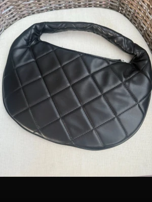 Svart quiltad handväska - Trendig svart handväska från Gina Tricot i quiltad design med mjukt konstläder. Väskan har en vadderad axelrem och dragkedja upptill. Insidan har ett fack med dragkedja för småsaker. Perfekt för dig som gillar stilrena accessoarer.