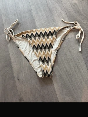 Randig bikiniunderdel med snörning - Säljer en bikiniunderdel med zigzag-randigt mönster i beige, svart och vitt. Modellen har knyt i sidorna för justerbar passform och är perfekt för sommaren och strandhäng. 