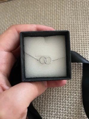 Silverfärgat armband - Snyggt armband i silverfärg med två sammanlänkade cirklar dekorerade med små glittrande stenar. Kedjan är tunn och elegant, perfekt för att lyfta vilken outfit som helst. Kommer i en svart presentask.