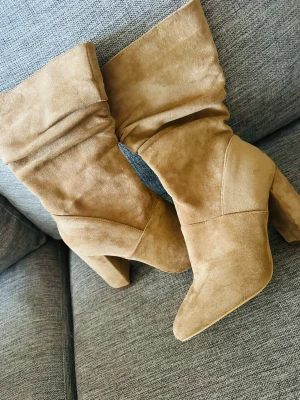 Oanvända beige stövletter med klack 🤎 - Jättefina beige stövletter i mockalook med hög, bred klack. Snygg och elegant modell med dekorativa veck och dragkedja på sidan.  Helt oanvända – aldrig använda. Sulorna är helt rena och skorna är i mycket fint skick, se bilder.  Säljer dem endast eftersom de inte kommer till användning. 🤎  Pris: 250 kr