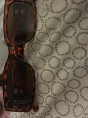 Rektangulära solglasögon leopardmönster - Säljer ett par trendiga solglasögon med rektangulära bågar i leopardmönster. Glasen är mörka och bågarna har en snygg brun ton. Perfekta för dig som vill sticka ut med en cool accessoar i sommar.
