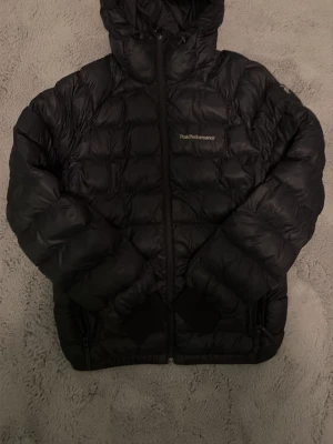 Svart dunjacka från Peak Performance - Snygg svart dunjacka från Peak Performance med huva och dragkedja framtill. Jackan har två fickor med dragkedja och quiltad design som ger en sportig look. Perfekt för kalla vinterdagar och riktigt varm tack vare dunfyllningen.
