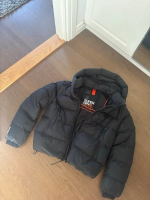 Svart pufferjacka från Superdry - Snygg svart pufferjacka från Superdry med huva och dragkedja framtill. Jackan har två sidofickor med dragkedja. Fodrad för extra värme och har elastiska muddar vid ärmsluten. Perfekt för kalla vinterdagar.