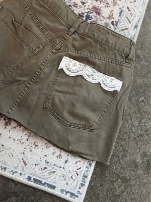 Olivgrön kortkjol med spetsdetalj - Snygg olivgrön kortkjol i bomull med två bakfickor och en vit spetsdetalj på ena fickan. Kjolen har en klassisk jeanskjols-look och passar perfekt till en avslappnad stil. Klippt