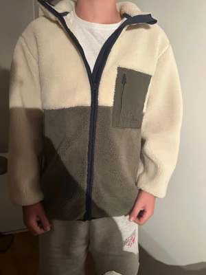 Beige och grå fleecejacka med dragkedja - Mysig fleecejacka i beige och grått med hel dragkedja framtill och hög krage. Jackan har en praktisk bröstficka med dragkedja i mörkgrått tyg och långa ärmar. Perfekt för kyliga dagar och enkel att matcha med andra plagg.