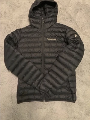 Svart dunjacka med huva Peak Performance - Säljer en svart dunjacka från Peak Performance med huva och dragkedja framtill. Jackan har quiltad design, två sidofickor och logga på bröstet samt ärmen. Perfekt för kalla vinterdagar och riktigt lätt att bära.