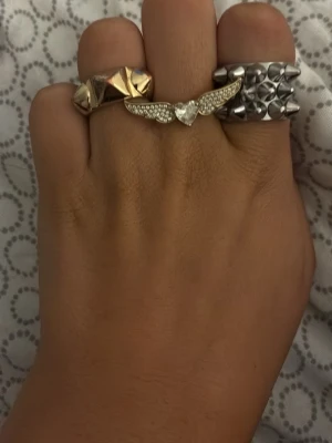Mixade ringar i guld och silver - Säljer ett set med sex ringar i olika stilar och material. Ringarna är i både guld- och silverfärg, vissa med stenar och detaljer som vingar, pärlor och geometriska former. Perfekt för dig som gillar att mixa och matcha accessoarer för en unik look.