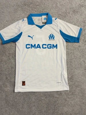Marseille matchtröja Puma vit/blå - Snygg vit matchtröja från Marseille med blå detaljer och klubbmärke på bröstet. Puma-logga och sponsortryck på framsidan. Tillverkad i lätt och ventilerande material, perfekt för fotboll eller träning.