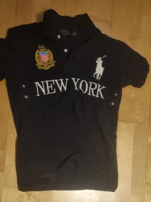 Chief keef polo blå new york - Marinblå pikétröja med stort vitt New York-tryck på bröstet, broderad logga och emblem samt siffran 5 på ryggen och ärmarna. Klassisk krage med knappar framtill. Materialet är bomull och modellen har korta ärmar.