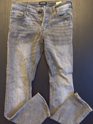 Grå tvättade jeans med knappar - Säljer ett par grå tvättade jeans med raka ben och knappgylf. Jeansen är i storklek W29 och L30 de har även fem fickor och bakfickor med diskret söm. Materialet är denim i bomull och passformen är slim Perfekta för en stilren outfit