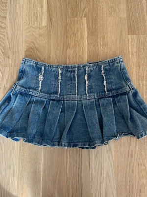 Kort  jeanskjol - Säljer en kort blå jeanskjol med plisserad nederdel och slitna detaljer framtill. Kjolen har dragkedja bak och en cool, ungdomlig vibe. Perfekt för dig som gillar denim och vill ha en edgy look.