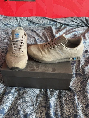 Adidas skor - Säljer ett par gråa Adidas sneakers med mesh-ovandel och klassiska tre ränder på sidan. Skorna har blå detaljer på tungan och vid sulan samt en svart yttersula. Lågt skaft och snörning framtill. Perfekta för dig som gillar stilren och sportig look. Har nog lite målarfärg p sig