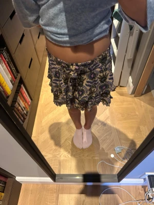 Blommig volangkjol med knyt - Supersöt kort kjol med blommigt mönster i beige, blått, grönt och lila. Kjolen har volanger och ett knytband i midjan som ger en extra detalj. Perfekt för dig som gillar boho-vibbar och vill ha något unikt i garderoben.