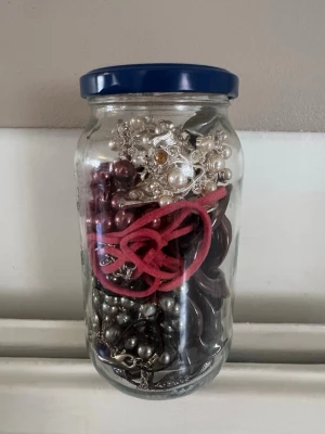 Blandade smycken i glasburk - En glasburk fylld med olika smycken som armband, halsband och ringar i blandade färger och material. Innehåller både metall, plast och textil. Perfekt för dig som vill ha unika accessoarer till din stil.