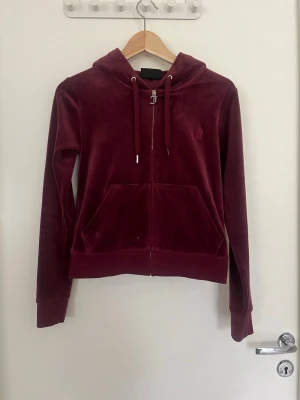 Vinrött velourset Juicy Couture - Mysigt loungewear-set i vinröd velour med matchande hoodie och byxor. Hoodien har dragkedja, fickor och dragsko i huvan. Byxorna har resår i midjan med dragsko och stora fickor framtill. Perfekt för chill dagar hemma eller när du vill vara extra bekväm. Två sömmar som släppt lite på fickorna fram på jackan som lätt går laga. 