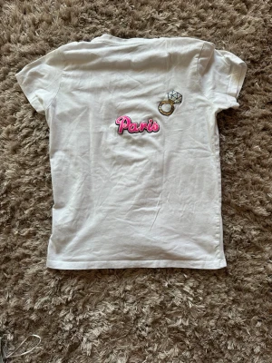 Vit t-shirt med Paris-tryck - Vit t-shirt i bomull med korta ärmar. På ryggen finns ett broderat motiv med texten 'Paris' i rosa och en ring med diamant. Enkel och clean design som passar till det mesta.
