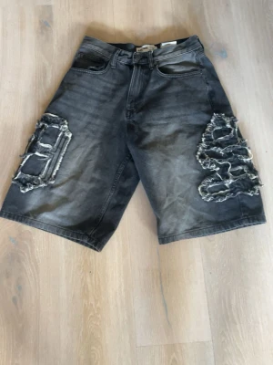 Svarta jeansshorts med slitningar - Säljer ett par svarta jeansshorts med grova slitningar och fransade detaljer på båda benen. Shortsen har fem fickor och en loose passform som ger en avslappnad streetwear-look. Perfekta för dig som gillar en edgy stil.
