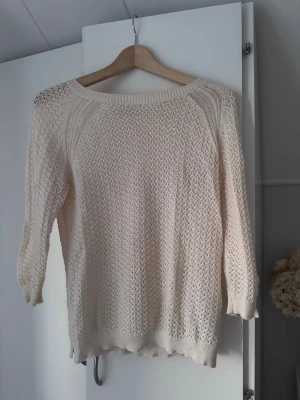 Krämvit stickad tröja - En krämvit stickad tröja med rund/boatneck halsringning och trekvartsärmar. Tröjan har ett luftigt, hålmönstrat stickat material och ribbade muddar vid ärmslut och nederkant. Perfekt för lager på lager och enkel att matcha till olika outfits.