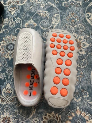 Nike mind grå  - Unika grå tofflor från Nike med futuristisk design. Ovandelen har perforerade hål för ventilation och sulan är täckt av orangea cirkulära pluppar för extra grepp. Tillverkade i ett mjukt, flexibelt material som känns bekvämt på foten. Perfekta för chill dagar hemma eller streetwear.