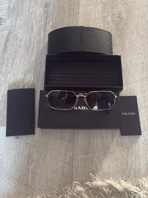 Prada solglasögon i silver - Snygga solglasögon från Prada med silverfärgad metallbåge och mörka glas. Modellen har en modern, kantig form och kommer med originaletui och tillbehör. Perfekt accessoar för dig som vill ha en exklusiv look. Köpta i Italien. Knappt använda. Köpt för 4000