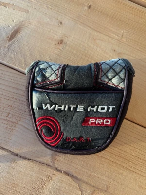 White Hot Pro putterfodral - Säljer ett svart och grått putterfodral med broderad text 'White Hot Pro' och röd spiral på framsidan. Fodralet har quiltade detaljer och stängs med kardborre. Passar till golfklubba och är gjort i konstläder och textil.