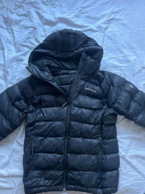 Svart dunjacka från Peak Performance - Snygg svart dunjacka från Peak Performance med huva och dragkedja. Jackan har quiltad design, två sidofickor med dragkedja och logga på bröstet.  Javkan har ett låtet hål vid en av fram fickorna som inte syns när jacka är på 