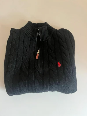 Svart kabelstickad tröja Polo Ralph Lauren - Kabelstickad svart tröja från Polo Ralph Lauren med halv dragkedja och ribbad krage. Tröjan har det klassiska röda Polo-logot broderat på bröstet och är långärmad. Perfekt för dig som gillar stilrena och tidlösa plagg.
