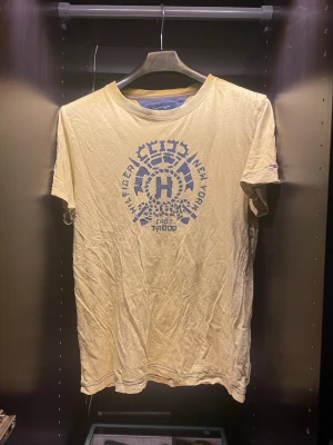 Beige t-shirt med blått tryck - Snygg beige t-shirt med blått grafiskt tryck på bröstet där det står 'Hilfiger New York East Troop'. Klassisk passform och korta ärmar. Perfekt till jeans eller shorts för en avslappnad look.