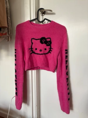 Hello Kitty rosa stickad crop tröja - Säljer en fluffig rosa stickad crop tröja med Hello Kitty-motiv framtill och svart Hello Kitty text längs båda ärmarna. Tröjan har rund halsringning och långa ärmar.