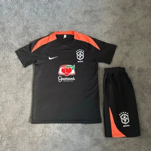 Svart Brasilien träningsset Nike storlek 164 - Snyggt svart träningsset från Nike med Brasilien-logga och Guaraná Antarctica-tryck. T-shirten har orange detaljer på axlarna och rund hals. Matchande shorts med orange inslag och Brasilien-emblem. Perfekt för fotbollsträning eller gymmet.En av fickornas dragkedja är trasig men fickan fungerar fortfarande 