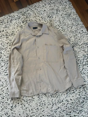 Beige skjorta från Zara - Stilren beige skjorta från Zara med klassisk krage och bröstficka. Skjortan har långa ärmar och knäppning framtill. Perfekt för en clean och enkel look, lätt att matcha med olika outfits.