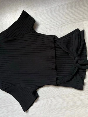 Svart ribbad topp med knyt - Säljer en svart ribbad topp med korta ärmar och rund hals. Toppen har en snygg knytning i ryggen som ger en unik look. Perfekt till jeans eller kjol för en trendig outfit.