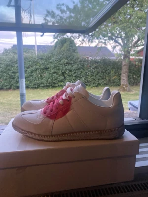 Maison Margiela gats - Maison Margiela sneakers i vitt skinn med en snygg rosa detalj på sidan och matchande rosa snörning. Klassisk siluett med beige sula och diskret branding under sulan. Perfekta för dig som vill ha något unikt och stilrent.