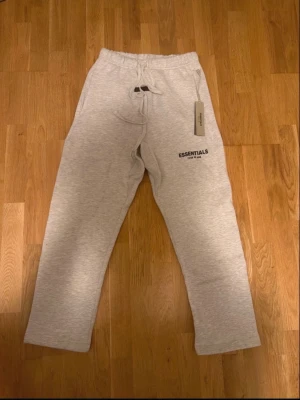 Gråa Essentials mjukisbyxor - Säljer ett par ljusgrå mjukisbyxor från Fear of God Essentials med snörning i midjan och logga tryckt på benet. Sköna och stilrena sweatpants i bomull, perfekta för chill dagar eller streetwear-look.