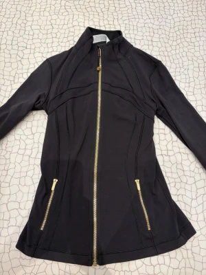 Lululemon Svart jacka med guldiga dragkedjor - Snygg svart jacka med figurnära passform och guldiga dragkedjor framtill och vid fickorna. Jackan har hög krage och långa ärmar, perfekt för en stilren look. Materialet känns mjukt och stretchigt, vilket gör den bekväm att bära. Endast använd 2 gånger. Nyskick. Priset går att diskutera. Skriv om du har nån fråga 