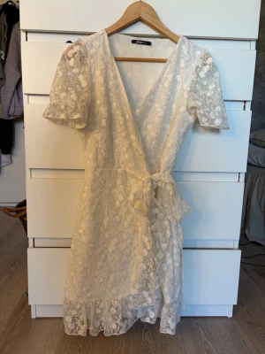 Vit spetsklänning med omlott och knyt - Romantisk vit kortklänning i spets med blommigt mönster. Klänningen har omlottmodell, knyt i midjan, volangkant nertill och korta puffärmar. Perfekt för sommarens alla tillfällen och ger en söt och feminin look.
