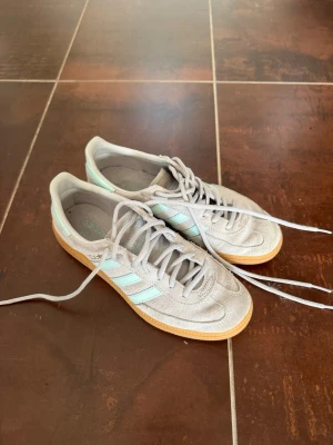 Adidas Spezial - Snygga sneakers från Adidas i grå mocka med klassiska mintgröna ränder på sidorna och matchande mintdetalj på hälen. Skorna har en låg profil och gummisula i brunt som ger en retrovibe. Perfekta för dig som gillar stilrena och sportiga sneakers.