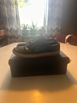 Svarta Saint Laurent sneakers - Säljer ett par svarta Saint Laurent sneakers med vit sula och lågt skaft. Skorna har klassisk snörning och Saint Laurent-logga på sidan. Tillverkade i Italien och levereras med originalkartong. Perfekta för dig som gillar stilrena och tidlösa sneakers.